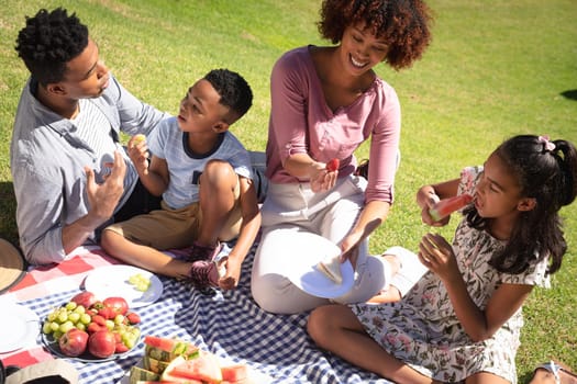 Happy african american couple with son and daughter outdoors, having picnic in sunny garden. family enjoying quality free time together.