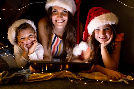 Happy caucasian mother, daughter and son wearing santa hats, using tablet at christmas time. family christmas time and festivity together at home.