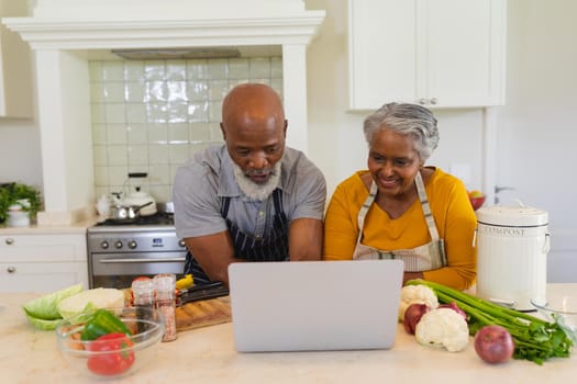Senior african american couple cooking together in kitchen using laptop. retreat, retirement and happy senior lifestyle concept.