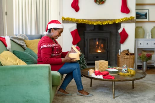 Happy african american senior woman in santa hat opening present at christmas time. retirement lifestyle and christmas festivities, celebrating at home.