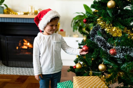 Happy caucasian boy wearing santa hat, decorating christmas tree at christmas time. childhood, chiristmas, festivity and tradition at home.