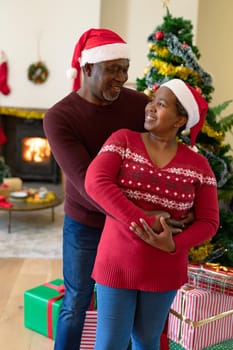Happy african american senior couple in santa hats dancing at home at christmas time. retirement lifestyle and christmas festivities, celebrating at home.