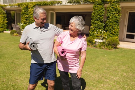 Senior caucasian couple embracing, holding yoga mats and smiling in sunny garden. retirement retreat and active senior lifestyle concept.