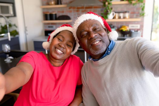 Happy african american senior couple taking selfie in kitchen at christmas time. retirement lifestyle, christmas festivities and communication technology.