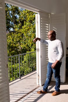 Thoughtful african american senior man standing at window and looking into distance. retirement lifestyle, spending time at home and garden.