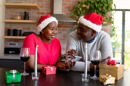 Happy african american senior couple in santa hats using smartphone in kitchen at christmas time. retirement lifestyle, christmas festivities and communication technology.