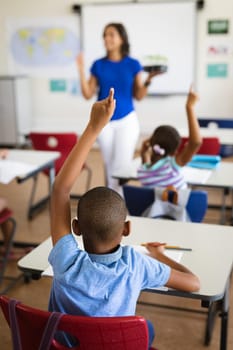 Rear view of african american boy raising his hands while sitting on his desk in the class at school. school and education concept