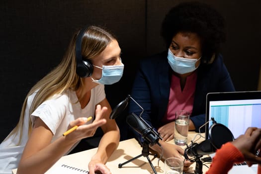 Diverse group of female business colleagues wearing masks in discussion speaking to microphones. recording podcast and business in a modern office during covid 19 coronavirus pandemic.