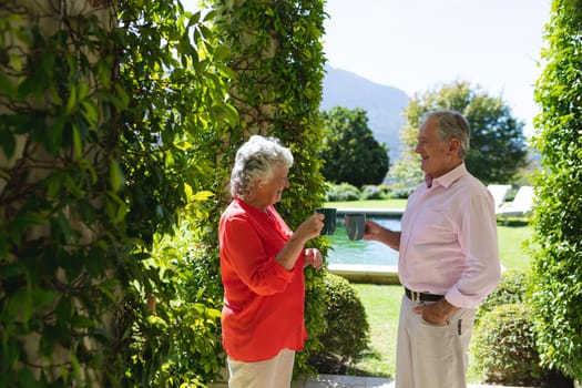 Senior caucasian couple smiling and holding mugs in sunny garden. retreat, retirement and happy senior lifestyle concept.