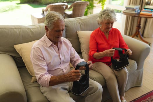 Senior caucasian couple sitting on sofa together holding vr headset in living room. retreat, retirement and happy senior lifestyle concept.