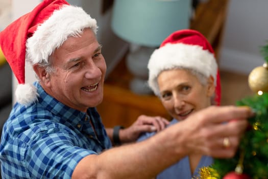 Happy caucasian senior couple wearing santa hat, decorating christmas tree. christmas, festivity and tradition at home.