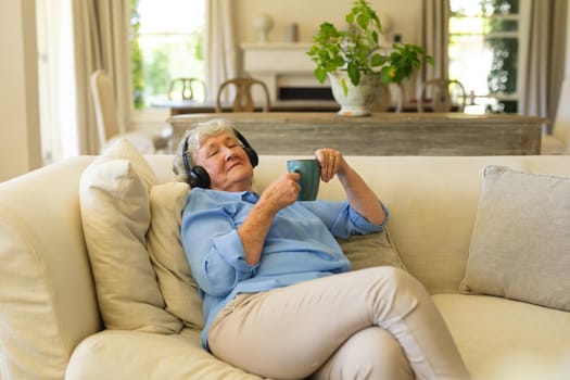Senior caucasian woman sitting on sofa wearing headphones with eyes closed. retreat, retirement and happy senior lifestyle concept.