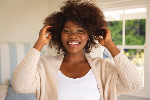 Portrait of smiling african american woman touching her hair sitting on bed at home. domestic lifestyle, enjoying leisure time at home.