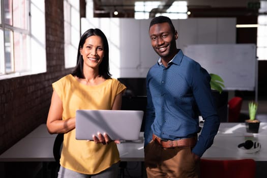 Portrait of smiling diverse male and female colleague standing in office looking at laptop together. working in business at a modern office.