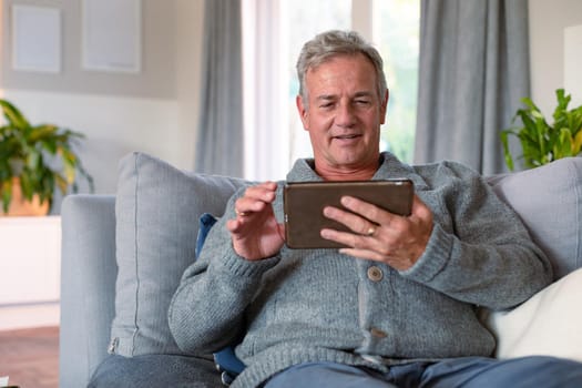 Happy caucasian senior man sitting on sofa and using tablet. active retirement lifestyle at home.