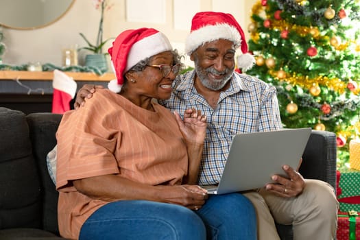 Happy african american couple wearing santa hats using laptop, having video call at christmas time. christmas, festivity and communication technology at home.