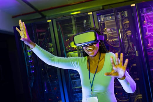 Happy african american female computer technician wearing vr headset and working in server room. digital information storage and communication network technology.