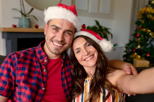 Happy caucasian couple wearing santa hats and taking selfie, christmas decorations in background. christmas, festivity and communication technology.