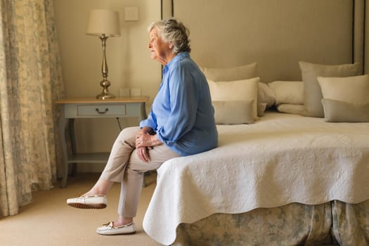 Senior caucasian woman sitting on bed and thinking in bedroom. retreat, retirement and senior lifestyle concept.