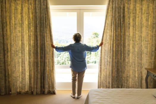 Senior caucasian woman standing next to window and widening courtains in bedroom. retreat, retirement and senior lifestyle concept.