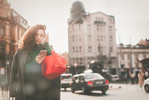 A woman on the street uses a mobile phone. online shopping. use of mobile applications. beautiful young woman with long curly dark hair in a casual coat, trendy green sweater and red handbag