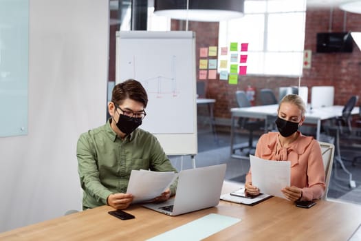 Diverse male and female colleague in face masks sitting at table with laptop reading paperwork. working in business at a modern office during coronavirus covid 19 pandemic.