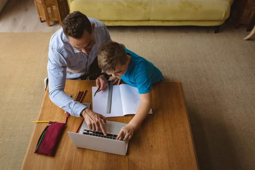 Overhead view of caucasian father using laptop to help his son with homework at home. home schooling and education concept
