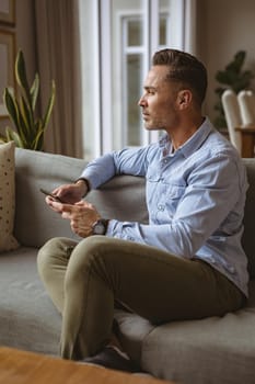 Thoughtful caucasian man using smartphone sitting on the couch at home. working from home during coronavirus covid-19 pandemic concept