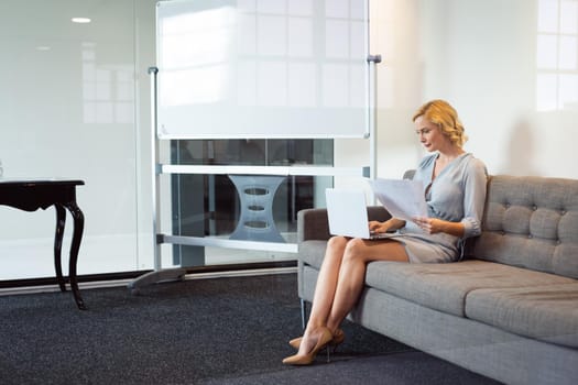 Caucasian businesswoman sitting in office lounge using laptop and holding paperwork. working in business at a modern office.