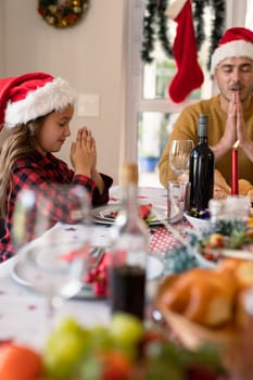 Caucasian daughter and father wearing santa hats praying at christmas table. family christmas time and festivity together at home.