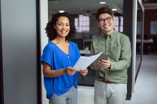 Portrait of smiling diverse male and female colleague with paperwork and tablet standing in office. working in business at a modern office.