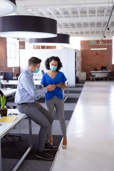 Diverse male and female colleague in face masks looking at tablet and discussing standing in office. working in business at a modern office during coronavirus covid 19 pandemic. .