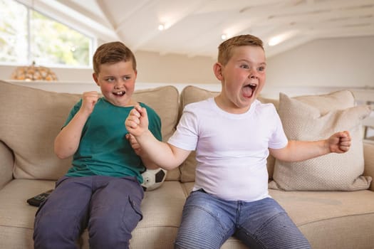 Two caucasian boys cheering while watching sports on television sitting on the couch at home. sports and entertainment concept