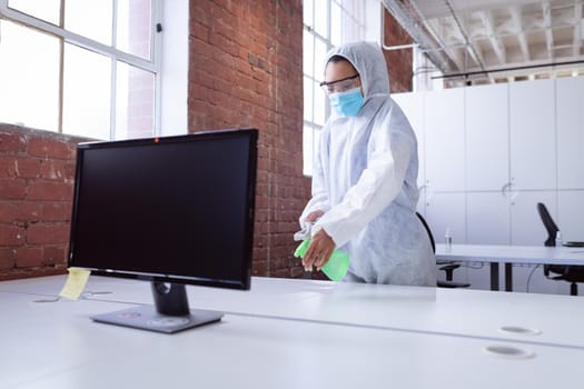 Cleaner wearing ppe suit, glasses and mask disinfecting office workspace, spraying desks. hygiene in business at a modern office during coronavirus covid 19 pandemic.