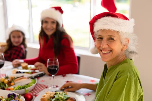 Happy grandmother, granddaughter and adult daughter wearing santa hats sitting at christmas table. family christmas time and festivity together at home.