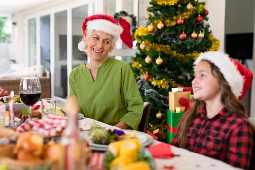 Happy caucasian grandmother and granddaughter wearing santa hats talking at christmas table. family christmas time and festivity together at home.