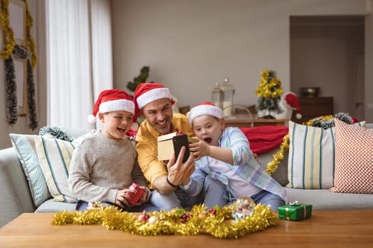 Caucasian father and two sons opening gift box during video call on digital tablet during christmas. social distancing during covid 19 pandemic at christmas time