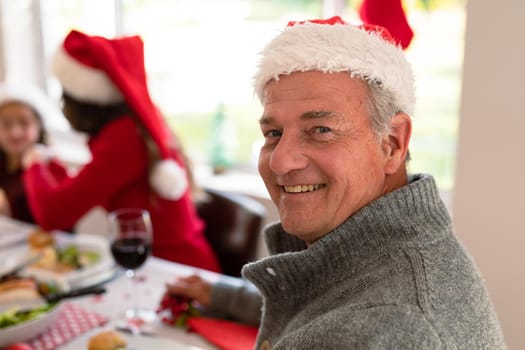 Happy senior caucasian man wearing santa hat looking at camera at christmas table. family christmas time and festivity together at home.