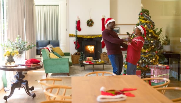 Happy african american senior couple in santa hats dancing at home at christmas time. retirement lifestyle and christmas festivities, celebrating at home.