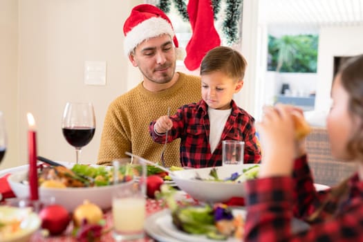 Happy caucasian father and son wearing santa hats sitting at christmas table. family christmas time and festivity together at home.