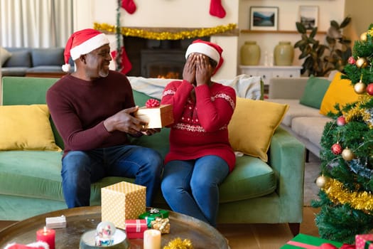 Happy african american senior couple in santa hats sharing presents at christmas time. retirement lifestyle and christmas festivities, celebrating at home.