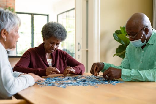 Two diverse senior women and african american male friend wearign face mask doing puzzles. socialising with friends at home.
