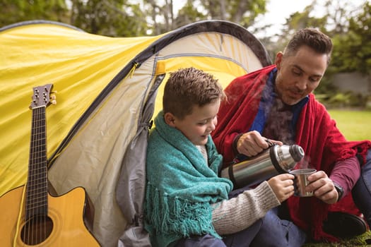 Caucasian father and pouring coffee in a cup for his son while sitting in a tent in the garden. fatherhood and love concept