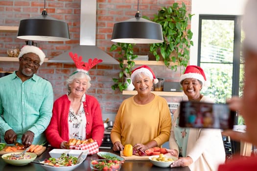 Group of happy diverse senior male and female friends in christmas hats taking selfie in kitchen. christmas festivities, celebrating at home with friends.
