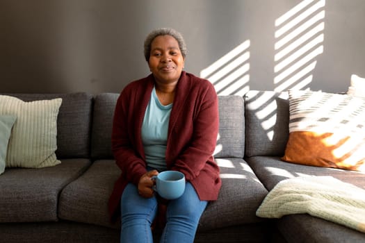 Relaxed african american senior woman sitting on sofa and drinking coffee. retirement lifestyle, spending time at home and garden.