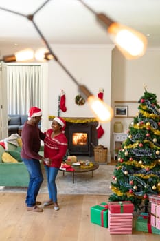 Happy african american senior couple in santa hats dancing at home at christmas time. retirement lifestyle and christmas festivities, celebrating at home.