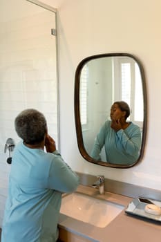 African american senior woman looking at mirror in bathroom. retirement lifestyle, spending time at home.