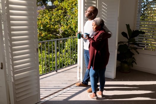 Relaxed african american senior couple drinking coffee standing on balcony in sun. retirement lifestyle, spending time at home and garden.