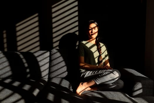 Serious asian woman sitting on sofa in ray of light at home. lifestyle, leisure and spending time at home.