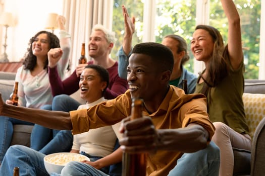 Group of happy diverse female and male friends watching tv and drinking beer together at home. socialising with friends at home.